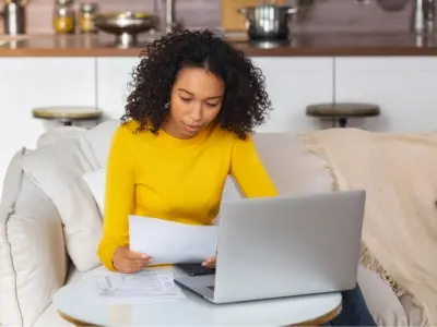 Woman in yellow sweater reviewing documents while working on a laptop at home, illustrating the importance of maintaining HVAC systems and managing utility bills.
