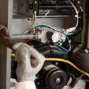 Hand in a glove repairing a mechanical component of an A-Plus air conditioning unit with visible tools and wires.