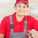 Smiling male HVAC technician in red shirt and gray overalls holding a wrench in workshop setting.