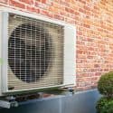 Air conditioning unit on red brick wall with circular fan grille and small shrub, featuring A-Plus HVAC solutions.