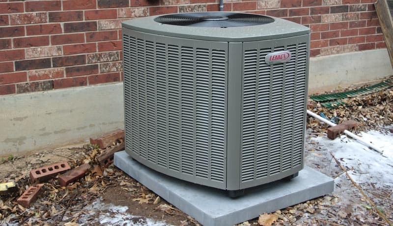 Air conditioning unit installed outside a residential building, featuring a Lennox brand logo, surrounded by leaves and bricks, illustrating HVAC solutions for home comfort.