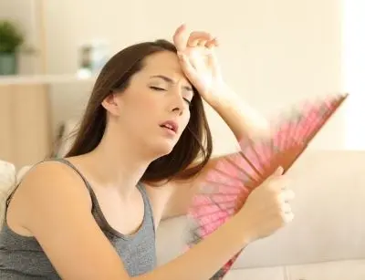 Woman fanning herself with a hand fan, appearing overheated and uncomfortable indoors, illustrating the need for effective air conditioning during warmer months.