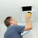 Man inspecting a ceiling vent with tools, promoting AC maintenance services from Atlantic Heating & Air Conditioning.