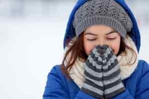 Woman in winter attire, wearing a blue coat and gray knitted hat, warming her hands in front of her face, surrounded by snow, emphasizing the need for boiler preparation in cold weather.