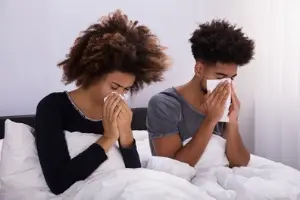 Couple sitting in bed with tissues, experiencing allergy symptoms, highlighting health impacts of dirty air conditioning.