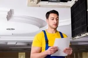HVAC technician in yellow shirt and blue overalls reviewing paperwork under an air conditioning unit, illustrating the essential role of HVAC professionals in maintaining comfort.