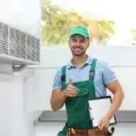 Smiling HVAC technician in green cap and overalls giving thumbs-up while holding clipboard beside air conditioning unit.