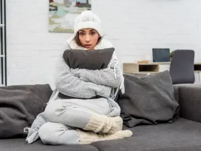 Woman sitting on a couch, bundled in warm clothing, holding a pillow, looking uncomfortable in a cold indoor setting, reflecting issues of uneven heating and humidity in homes.