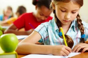 Young girl writing with a pencil at a school desk, surrounded by classmates, with a green apple in the foreground, emphasizing back-to-school preparations and the importance of organization for parents.