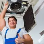 Smiling male technician in blue overalls gives thumbs up next to an indoor air conditioning unit during maintenance.