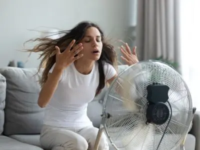 Woman sitting on a couch, frustrated and fanning herself in front of a fan, illustrating inadequate cooling power and discomfort during summer heat.