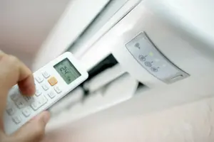 Hand holding remote control aimed at a wall-mounted air conditioning unit, illustrating HVAC system operation and maintenance.