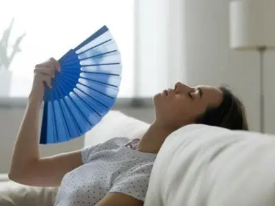 Woman using a blue fan to cool off indoors, illustrating discomfort from inadequate air conditioning in a home setting.