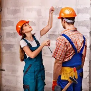 Two construction workers in hard hats discussing a property inspection, with one measuring or checking wiring in a home setting.