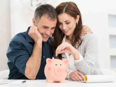 Couple happily saving money by depositing coins into a pink piggy bank, symbolizing financial planning and savings for home maintenance.