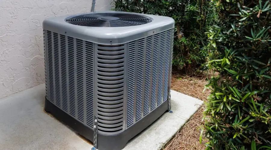 Gray air conditioning unit outside North Point Air Conditioning building on concrete slab with green bushes nearby.