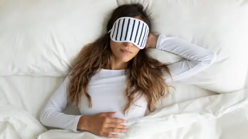 Woman wearing a sleep mask lying comfortably in bed, illustrating the importance of optimal bedroom conditions for restful sleep.