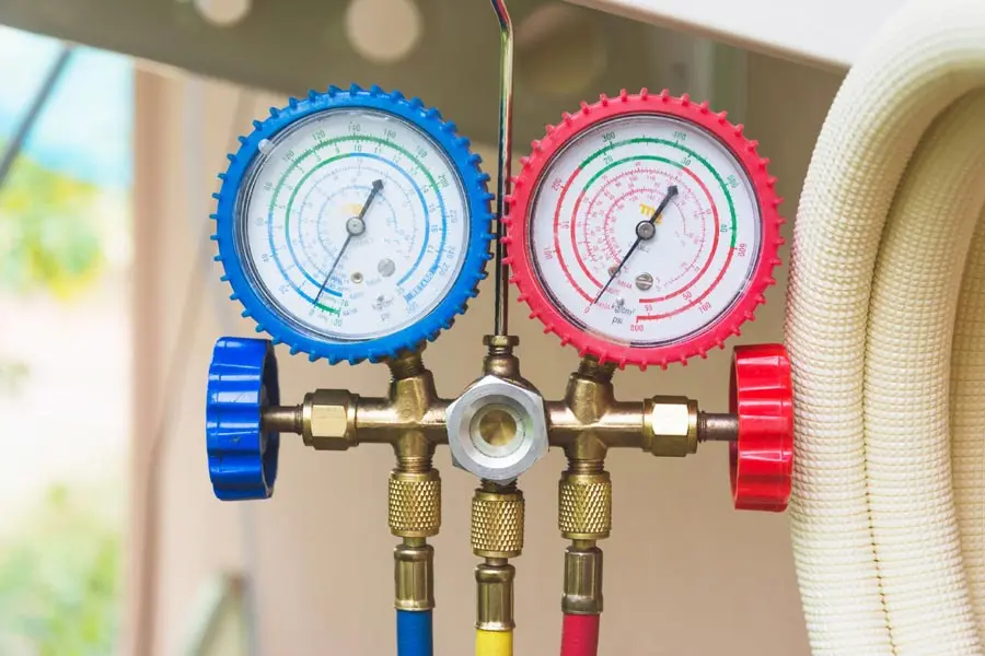 HVAC pressure gauges with blue and red dials, used for air conditioning maintenance, alongside hoses in a home setting, reflecting the importance of system upkeep for seasonal transitions.