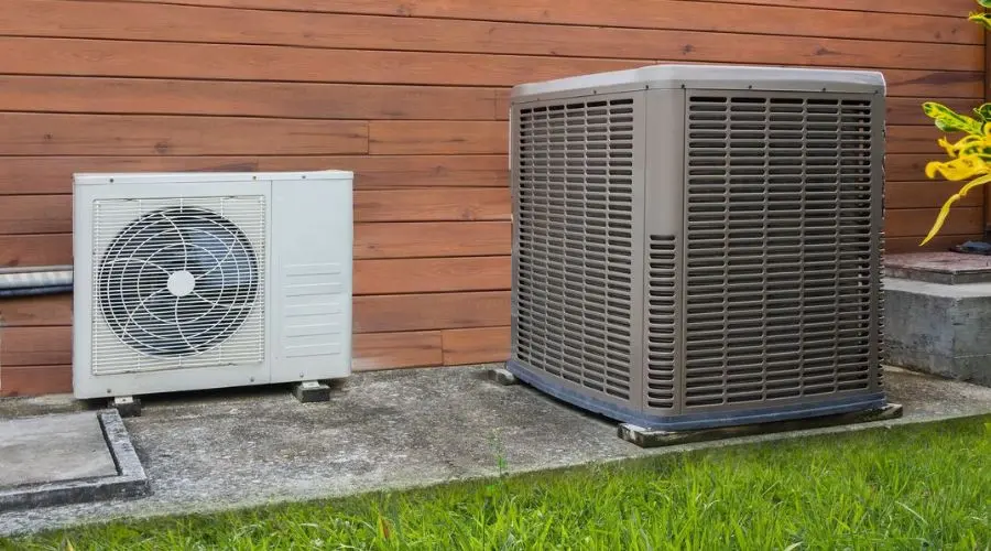 Air conditioning units on a concrete slab, featuring a smaller white unit and a larger dark unit, surrounded by grass.