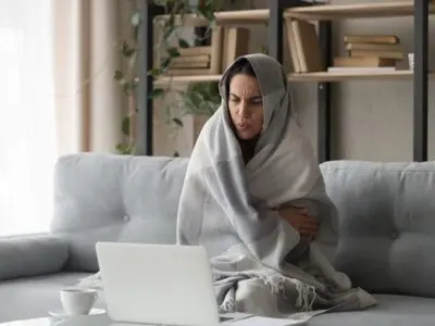 Woman wrapped in a blanket sitting on a couch, looking cold and uncomfortable while using a laptop, illustrating the importance of effective heating solutions like heat pumps for home comfort.
