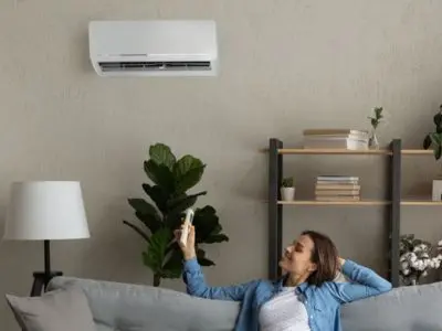 Ductless mini-split air conditioning unit installed on wall, woman relaxing on couch with remote control, indoor living space with plants and bookshelves, demonstrating home cooling solutions.