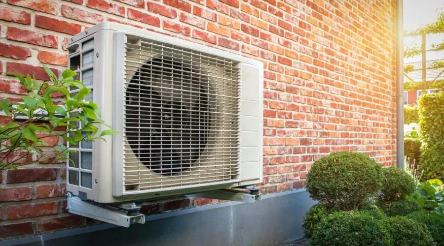 Air conditioning unit mounted on a brick wall with green shrubs in sunlight, showcasing North Point HVAC services.