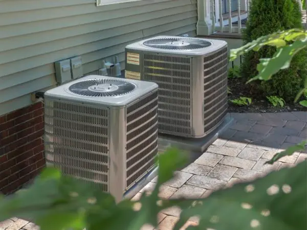 Two air conditioning units positioned beside a house, surrounded by greenery and paving stones, illustrating HVAC systems relevant to soundproofing solutions for improved home comfort.