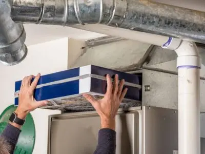Person replacing a furnace filter to prevent overheating, with visible ductwork and furnace unit in background.