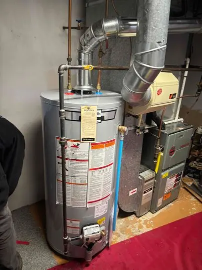 Newly installed residential hot water heater with connected ventilation and copper piping. A Postma installation tag is attached to the unit. Furnace system is visible beside the water heater in a clean utility room.