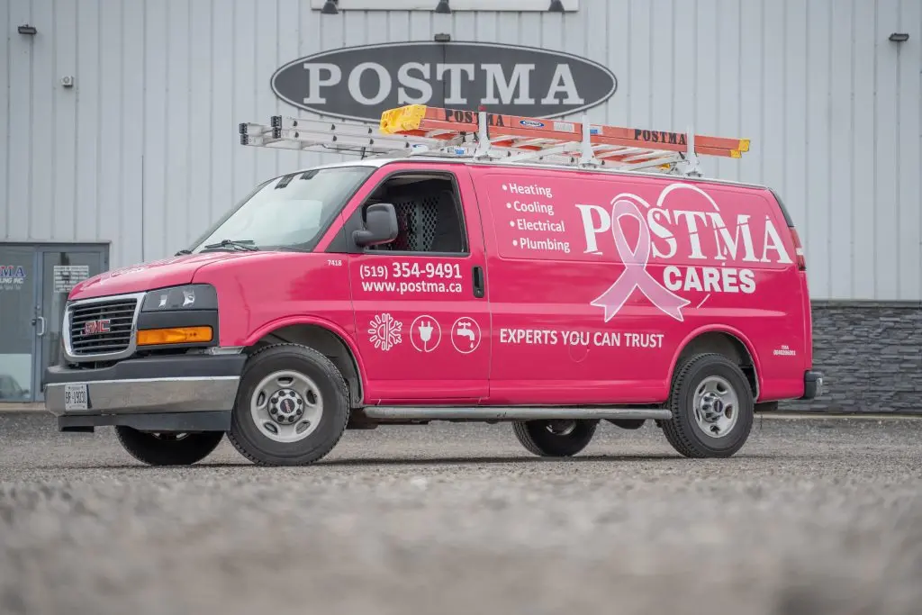 The image shows a bright pink Postma Heating, Cooling, Electrical, and Plumbing service van parked in front of the Postma building. The van features a large ribbon symbol in support of cancer awareness, along with the text "POSTMA CARES" prominently displayed. The company’s services—heating, cooling, electrical, and plumbing—are listed on the side of the van, accompanied by service icons. The company’s contact information, including the phone number (519) 354-9491 and the website www.postma.ca, are also clearly visible. The phrase "Experts You Can Trust" is displayed beneath the service listings, emphasizing the company’s reliability and commitment to the community.