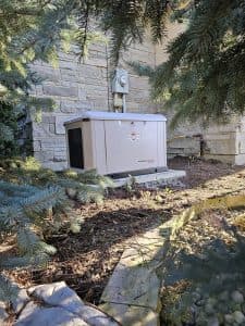 Home generator installed, partially surrounded by evergreen trees. The generator, a Briggs & Stratton PowerProtect model, is securely connected to the home's electrical system, demonstrating professional generator installation. The setup is designed to provide reliable backup power, highlighting the importance of generator maintenance to ensure consistent operation during power outages.