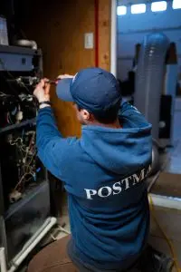 Postma HVAC technician performing furnace maintenance, using tools to service internal components of a residential heating unit inside a utility room.
