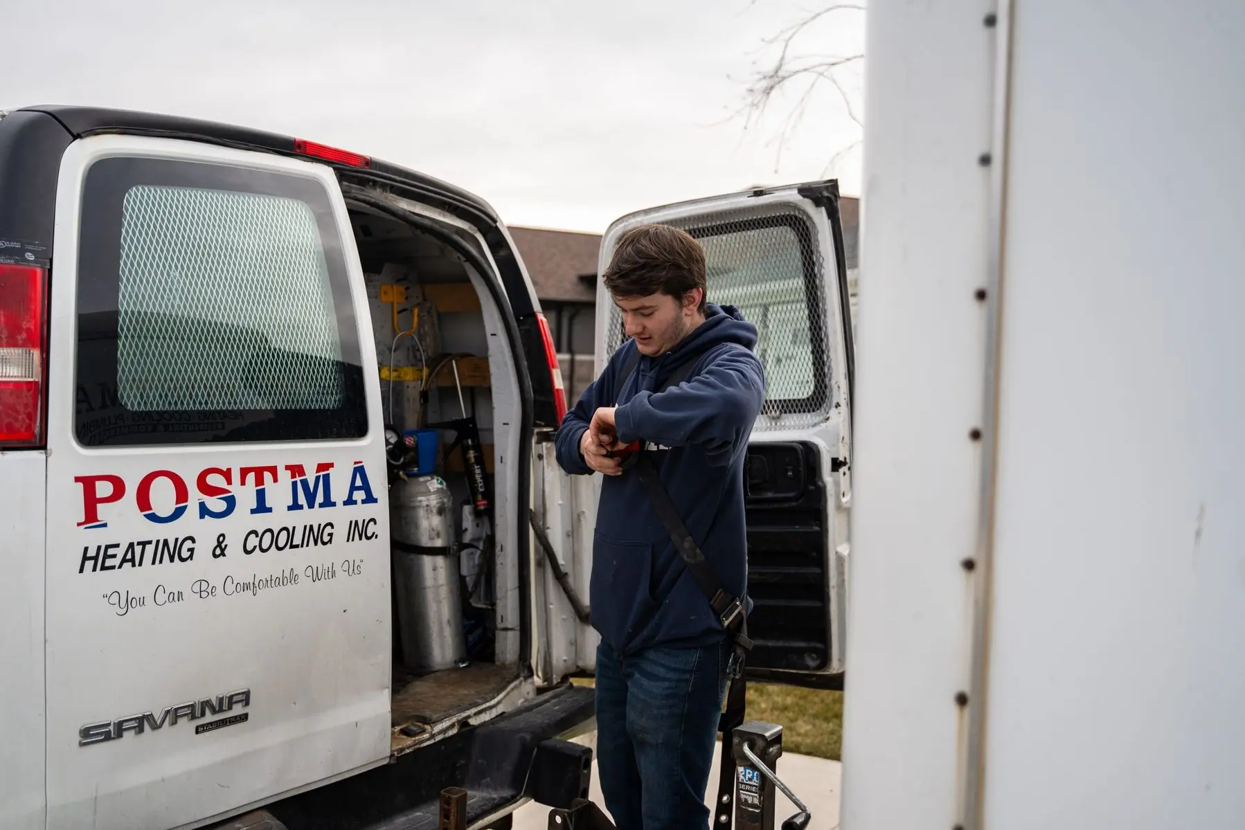 Postma Heating & Cooling technician in Chatham preparing for a job, standing beside an open service van with tools inside. The van displays the company name and slogan, 'You Can Be Comfortable With Us.