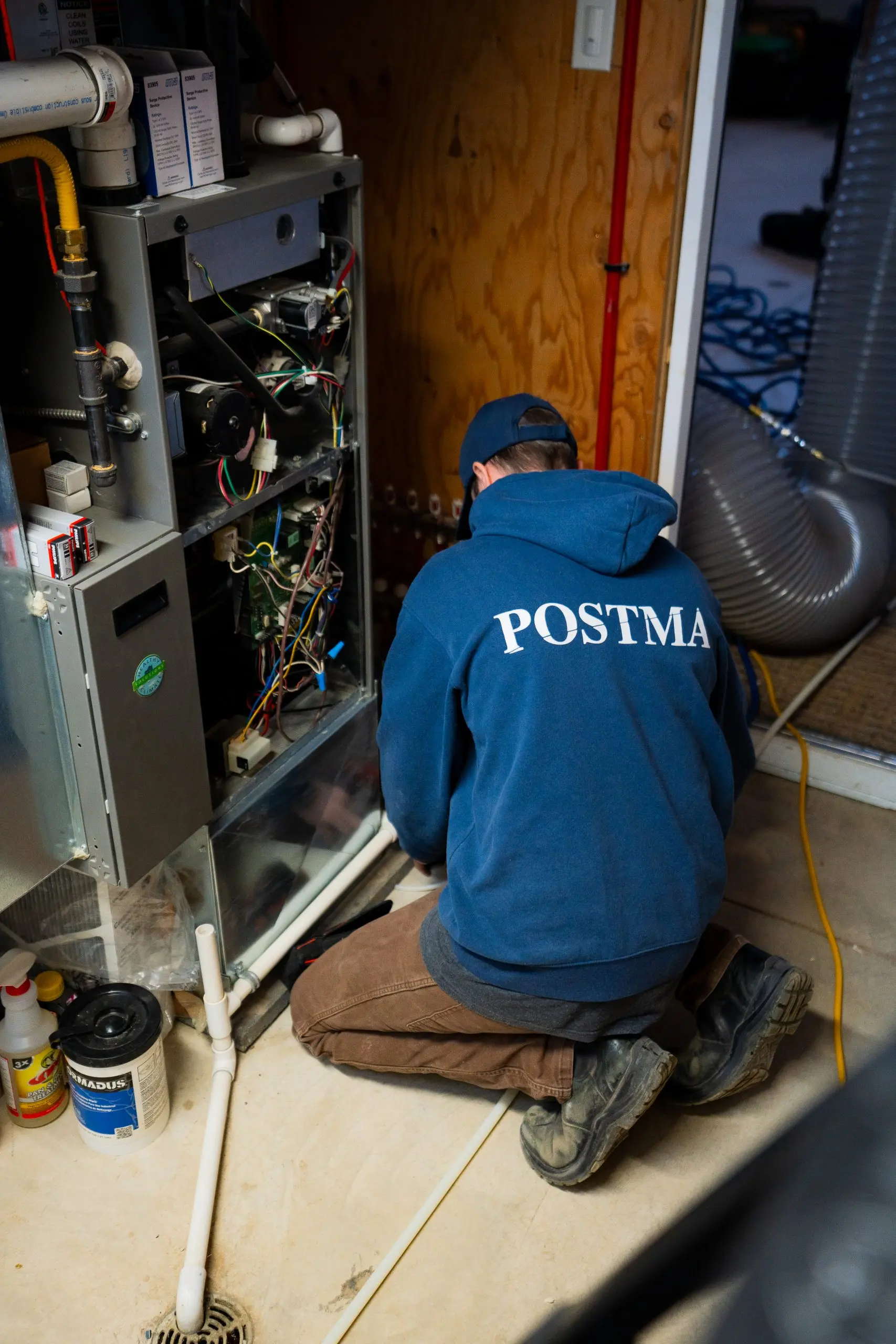 Postma technician kneeling while performing maintenance on a residential furnace, with the unit's front panel removed to access internal wiring and components.