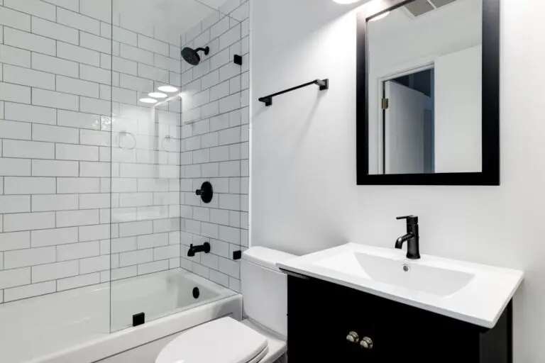 A small modern bathroom with a dark vanity, mirror frame, and hardware. White subway tiles line the bathtub and shower with black faucets. New bathroom fixtures.