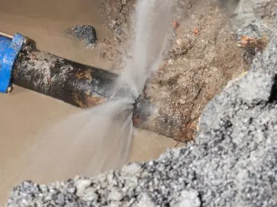 Burst water pipe leaking, surrounded by muddy water and gravel, indicating plumbing issues related to water leaks and low pressure.