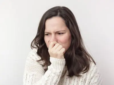 Woman in a white sweater grimacing and holding her nose, indicating a reaction to an unpleasant odor, relevant to unusual smells from heating systems.