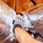 Gloved hand inspecting silver foil ductwork among wood beams for Air Works Heating & Air HVAC services.