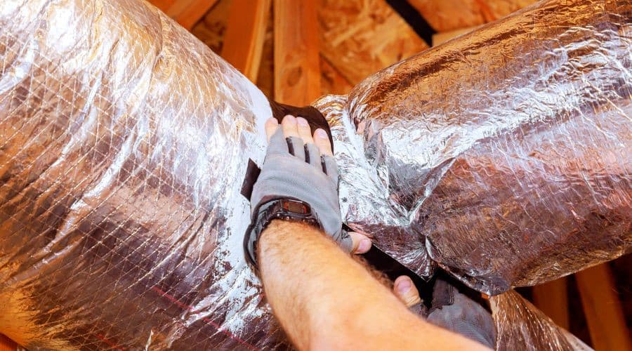 A gloved hand inspecting shiny metallic duct insulation near wooden beams for HVAC maintenance.