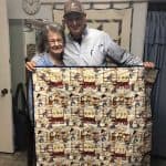 Elderly woman and man showcasing a quilt in a cozy setting, with a wall clock in the background.