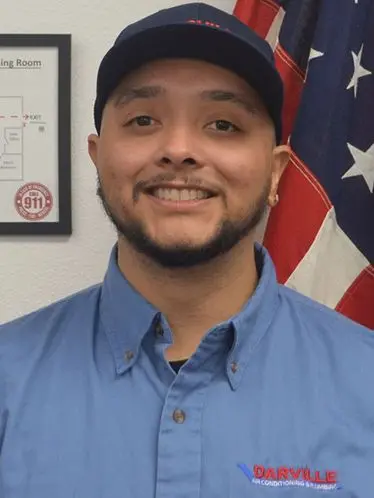 Antonio Hernandez, plumbing technician at The Darville Company, smiling in a blue shirt with company logo, American flag in background.