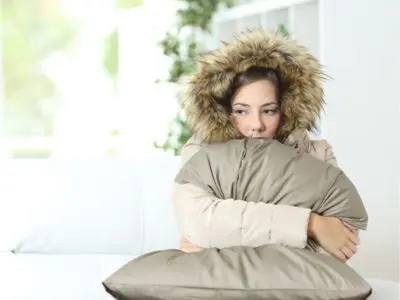 Woman in a warm coat hugging a pillow, conveying discomfort from cold indoor temperatures, emphasizing the importance of effective heating solutions.