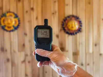 Hand holding an indoor air quality monitor displaying humidity and temperature levels against a wooden wall with decorative sun masks, illustrating the importance of air quality assessment for home comfort and HVAC maintenance.