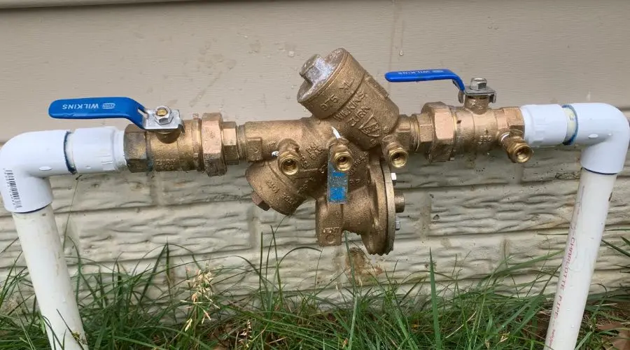 Backflow preventer valve with blue handles connected to PVC pipes, situated near a home’s foundation, illustrating essential plumbing safety for municipal water systems.