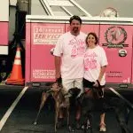 Couple in matching pink t-shirts with greyhounds by a pink service vehicle, showcasing pet charity support.