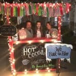 Hot cocoa stand with festive decorations, featuring two adults and a child enjoying drinks during a winter celebration.