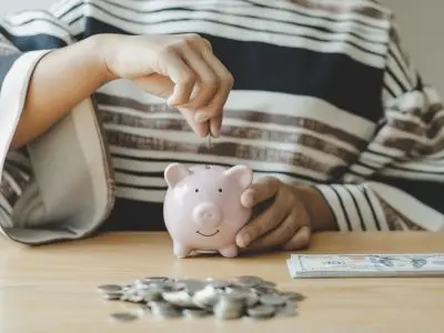 Person depositing a coin into a pink piggy bank, surrounded by scattered coins and a stack of bills, representing savings and financial management.
