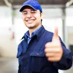 Smiling technician in blue cap and uniform gives thumbs-up in HVAC workshop, showcasing Chad's AC Direct services.