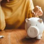 A person saving money by inserting a coin into a white piggy bank on a wooden table, emphasizing financial responsibility.