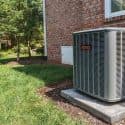 Air conditioning unit in residential backyard with brick wall, grass, and trees, showcasing Chad's HVAC services expertise.